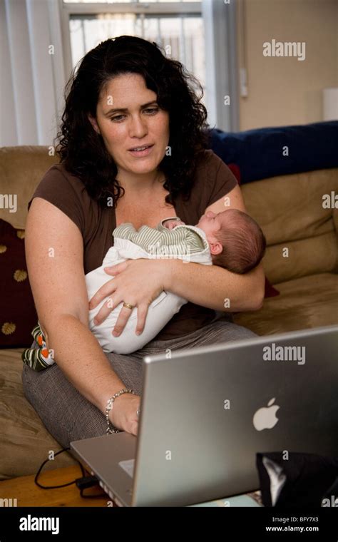 New mother holds her newborn baby while working on an Apple laptop computer at home in her ...