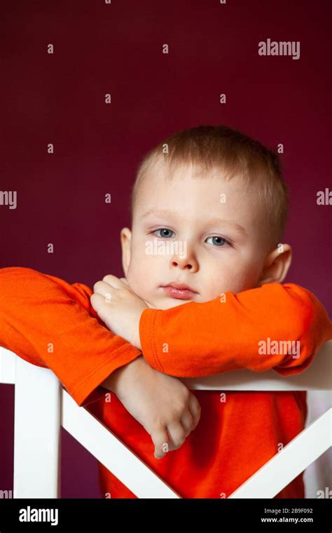 Thoughtful little blond boy sitting on a white kitchen chair as he stares at the camera. Cute ...