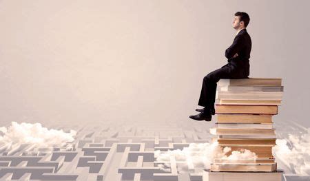 A serious businessman in suit sitting on a pile of giant books in front of a grey wall with ...