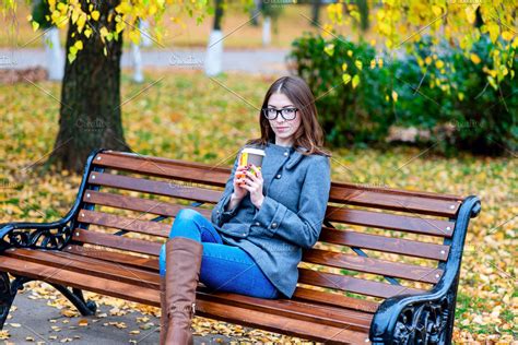 Beautiful young woman sitting on a bench drinking coffee or hot tea in the spring autumn coat ...