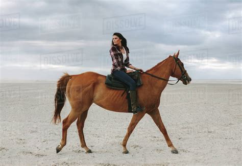 Woman horse ride on beach at sunset and glancing back. Beautiful female riding a brown horse in ...