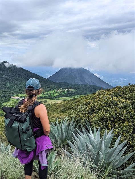 a woman standing on top of a lush green hillside with mountains in the ...