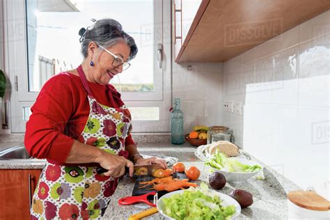 Side view content grandma in apron cutting tomatoes while cooking dinner in contemporary kitchen ...