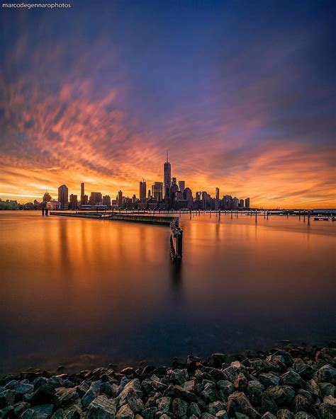 a city skyline is seen at sunset over the water with rocks in front of it