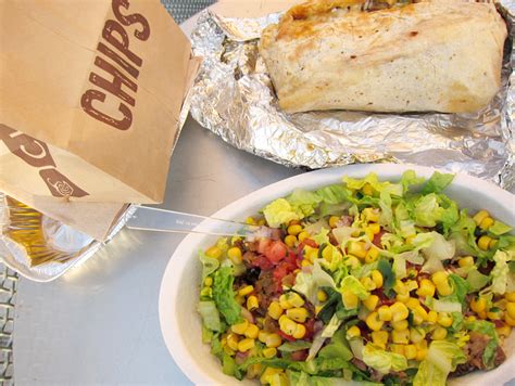 a bowl of salad next to a burrito and some chips on a table with a paper bag