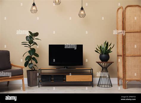 Modern TV on cabinet, armchair and green plants near beige wall in room ...