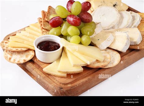 A platter of different cheese with grapes, biscuits, and raspberry jam on a wooden board ...