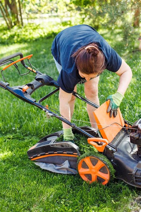 Vertical Shot of Brown Haired Female Gardener Raking Fresh Grass Out Lawn Mower or Grass Cutter ...