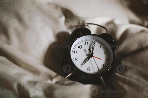 Classic black alarm clock showing seven o'clock with a bed in the ...