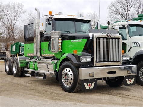 two green and white semi trucks parked in a parking lot with bare trees behind them