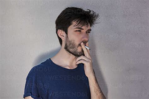 Young man with beard smoking cigarette in front of cement wall stock photo