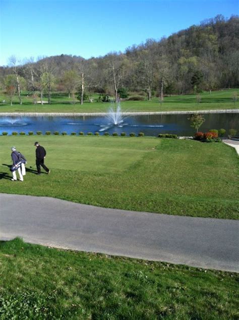 two people sitting on benches in the grass near a pond and park with water spouting from it