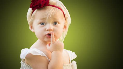 Cute Little Girl Baby Is Keeping Finger On Lips In Green Blur Background Wearing White Dress And ...