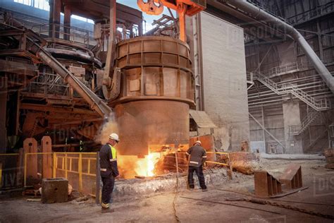 Steelworkers testing pour of molten steel from ladle in steelworks - Stock Photo - Dissolve