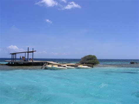 two people standing on a dock in the middle of clear blue water near an island