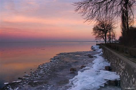 the sun is setting over the water and ice on the shore near some trees with no leaves