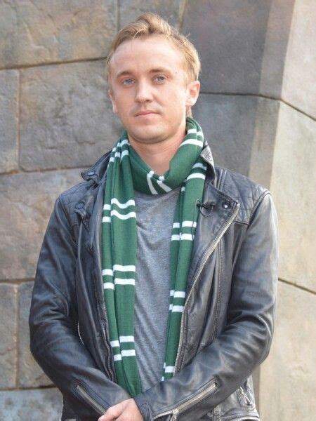 a man wearing a green scarf standing next to a stone wall with a black leather jacket on