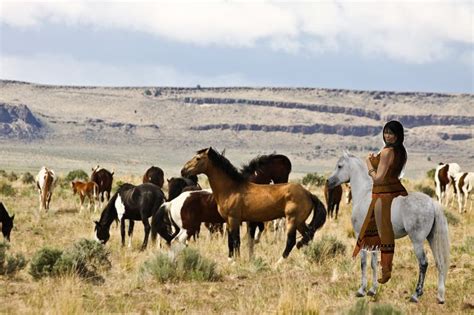 Native American Woman riding her grayish white horse while watching over a herd of wild mustangs ...