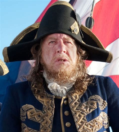 a man with long hair wearing a black hat and blue coat standing in front of an american flag