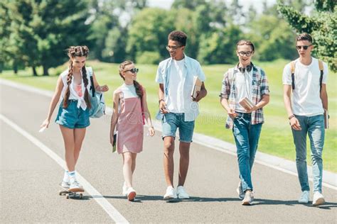 Multiethnic Group of Teenagers with Books and Skateboard Talking and Walking Together in Park ...