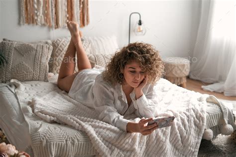 Young barefoot woman lying on cozy bed and watching movie on smartphone Stock Photo by ...