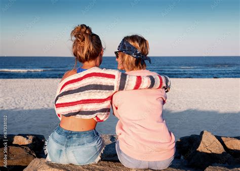 Two young girls, best friends sitting together on the beach at sunset ...