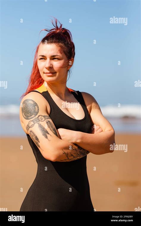 Self assured female athlete with crossed arms looking away on sandy ocean beach in sunlight ...