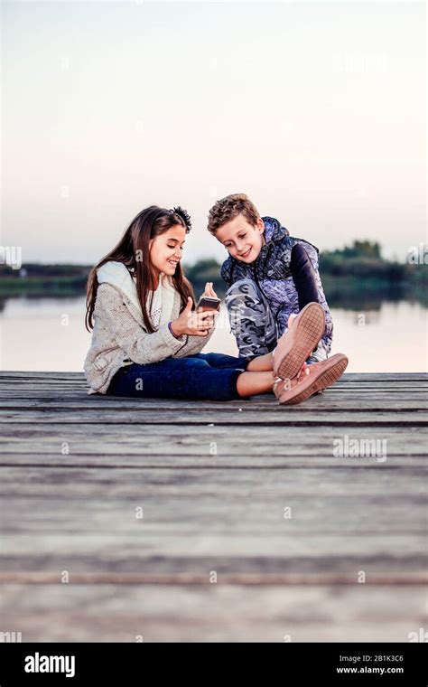 Two young cute little friends, boy and girl having fun wih smart phone sitting by the lake in ...