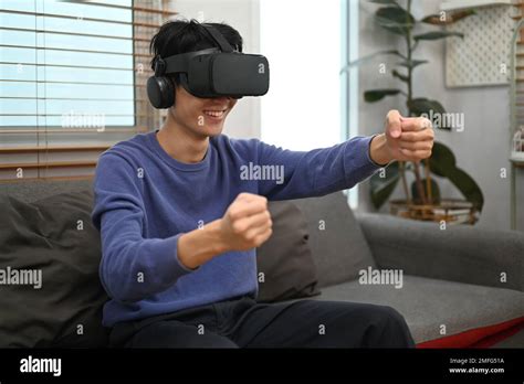 Cheery Asian man in VR headset holding imaginary steering wheel of virtual car, playing a ...