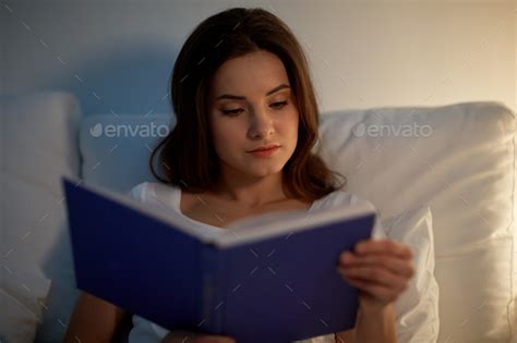 young woman reading book in bed at night home Stock Photo by dolgachov