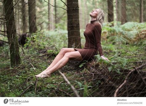 Young blonde woman sitting barefoot on fallen tree trunk in forest looking at sky - a Royalty ...