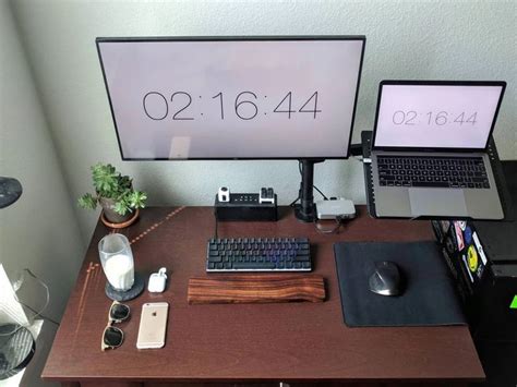 a desk with a laptop, keyboard and mouse on it next to a monitor displaying the time