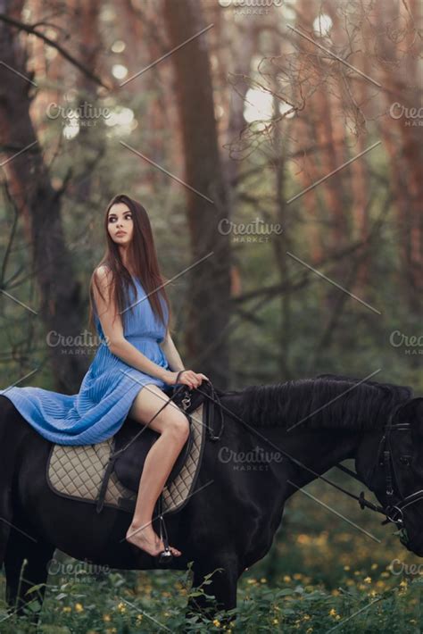 Young beautiful brunette girl in blue dress ride on the black horse in forest Young And ...