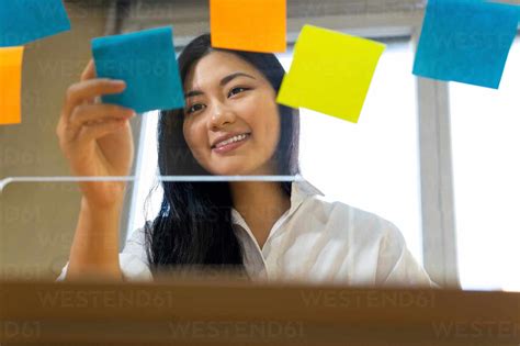 Young glad ethnic female entrepreneur arranging colorful paper stickers on transparent surface ...