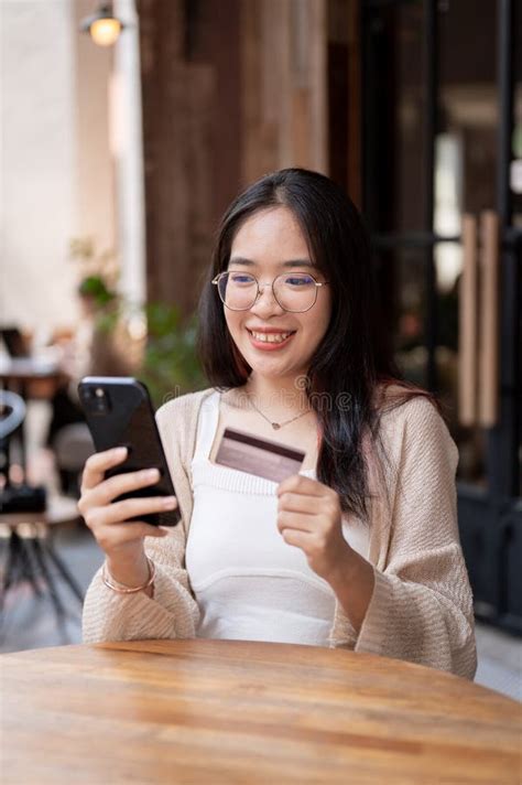 A Happy Asian Woman Sits at an Outdoor Table of a Cafe, Holding Her Credit Card and Smartphone ...