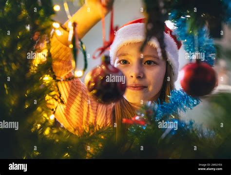 Jolie petite fille en chapeau de père Noël rouge accrochant une boule de décoration d'arbre ...