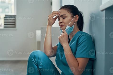 Stressed young female nurse in protective face mask crying while working in the hospital ...