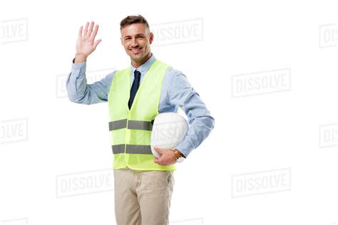 Smiling engineer holding helmet, waving and looking at camera isolated ...