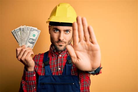 Young Builder Man Wearing Safety Helmet Holding Dollars As Payment Over Yellow Background with ...