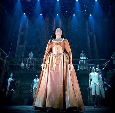 a woman in an orange dress standing on stage with other people behind her and lights above her