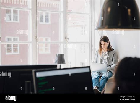 Focused Young Female Programmer Using Laptop While Sitting By Window In