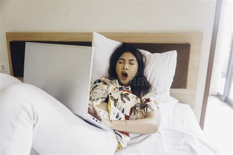 A Sleepy Young Woman Yawning While Working From The Bed Stock Photo