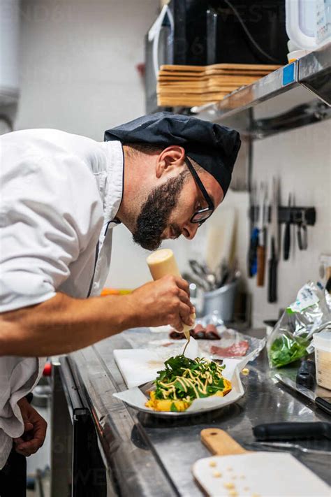 Side View Of Busy Male Chef In Uniform Adding Sauce On Top Of Dish With