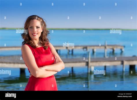 Brunette Woman Dress In Red Smiling Relaxed On A Blue Lake Jetty Stock