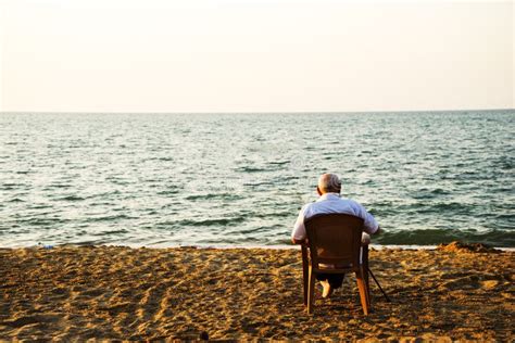 Old Man Sitting On A Chair On The Beach And Looking At The Sea Stock