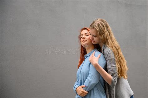 Two Happy Girlfriends Are Hugging On The Background Of A Gray Wall