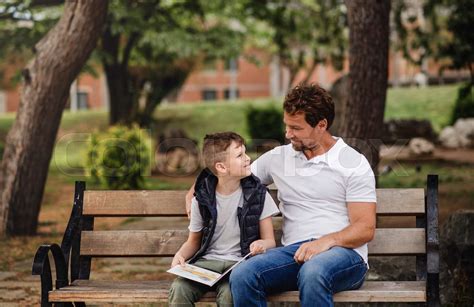 Father With Son Sitting On Bench Outdoors In Town Reading A Book