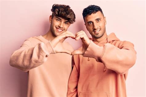 Joven pareja gay con ropa informal sonriendo enamorada haciendo forma de símbolo de corazón con