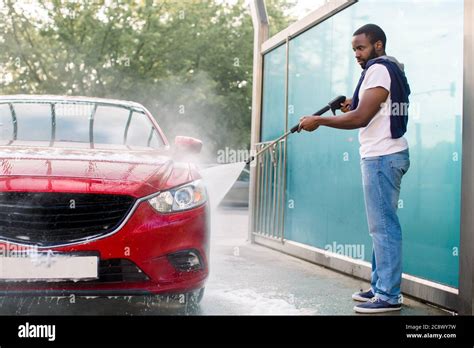 Handsome African guy in t-shirt and jeans washing his red luxury car ...