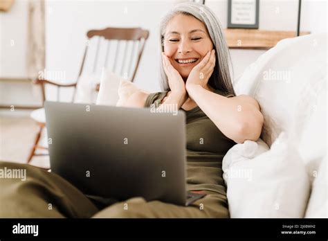 Mature Asian Woman Laughing And Working With Laptop While Sitting On
